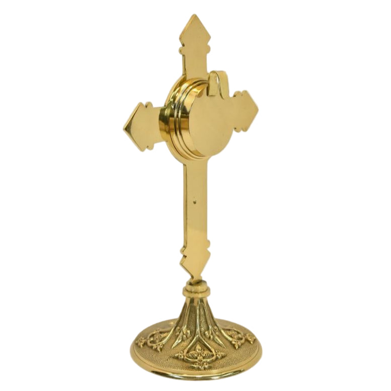 A polished brass cross-shaped reliquary with a round relic compartment and an ornate base. Side view showing hook on the back for veneration.